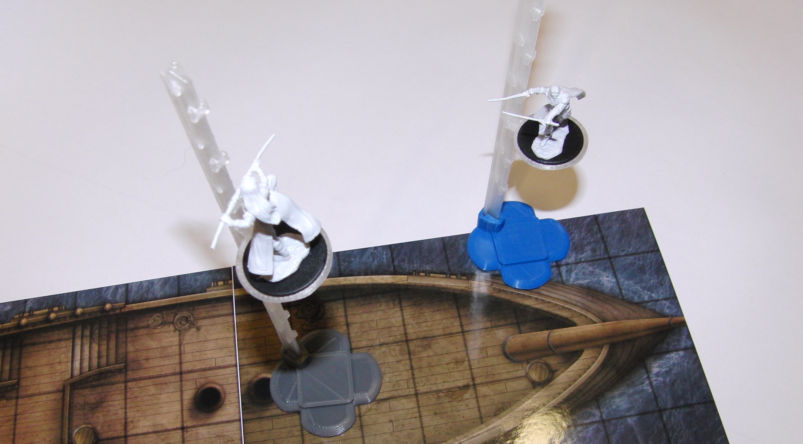 A tabletop with two minis atop a boat mat grid.
                        The two minis are on flying stands.  One flying stand
                        has a grey base, while the other flying stand has a
                        blue base.