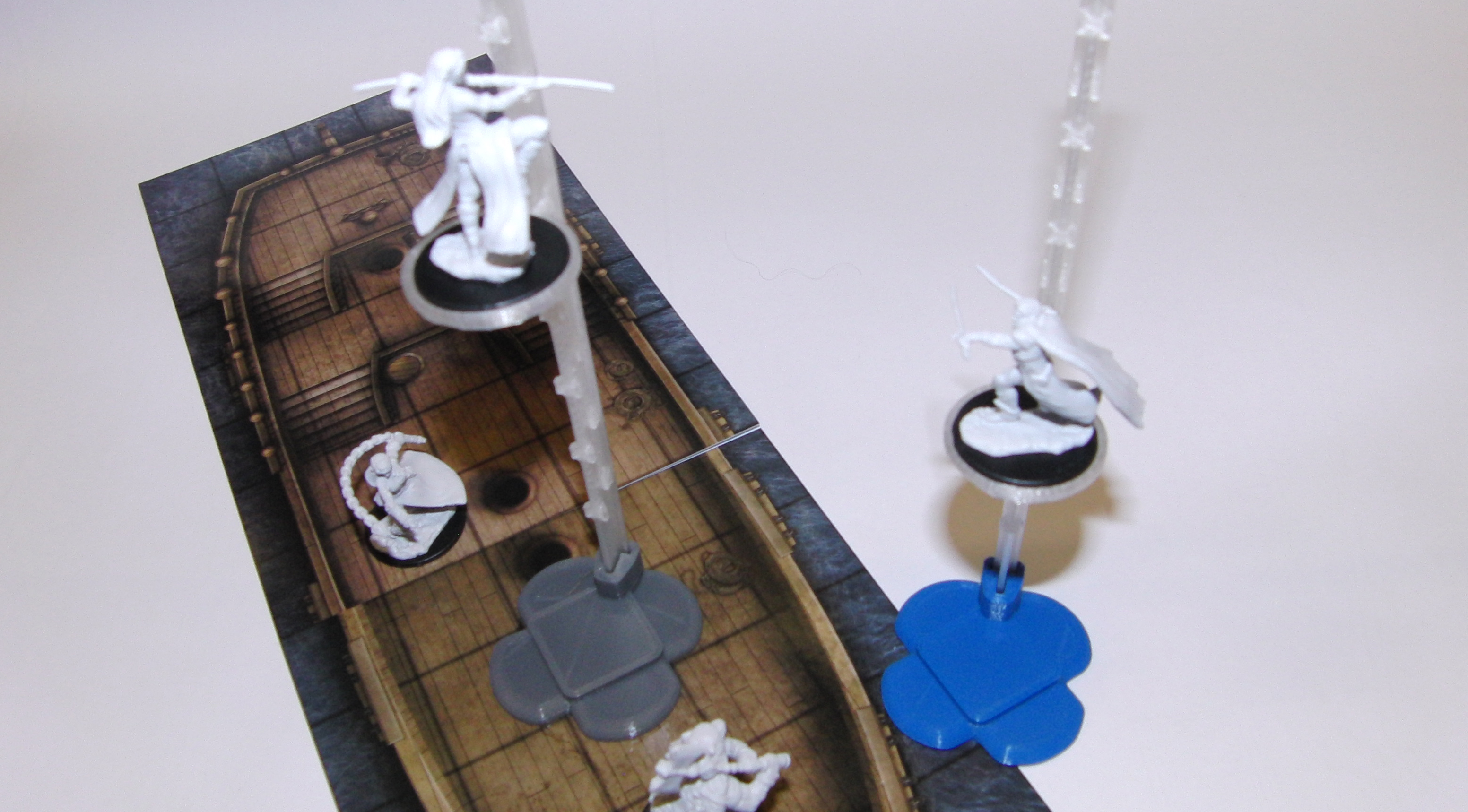 A tabletop with four minis atop a boat mat grid.
                        Two minis are on flying stands.  One flying stand has
                        a grey base, while the other flying stand has a blue
                        base.  The other two minis are not on flying stands.
                        They are on the boat.